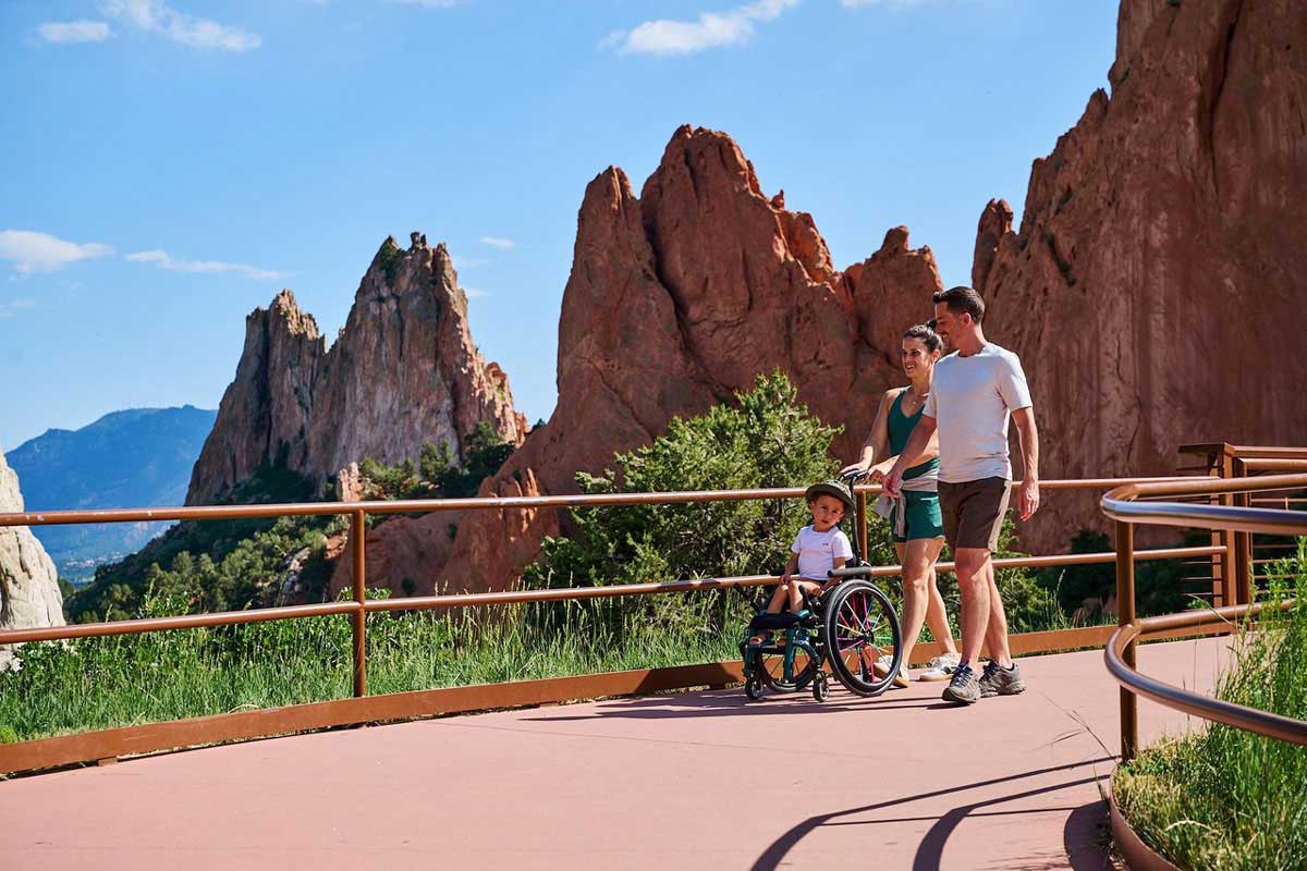 A family of two parents and a child in a wheelchair explore the red-rock spires of Colorado Springs' Garden of the Gods on a paved path. 