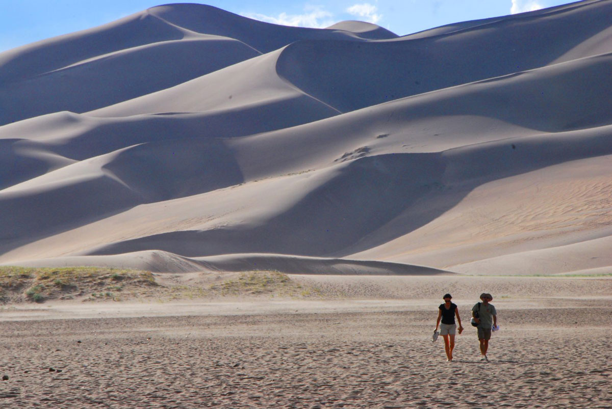 Two people walk through the sand below giant dunes at Great Sand Dunes National Park & Preserve