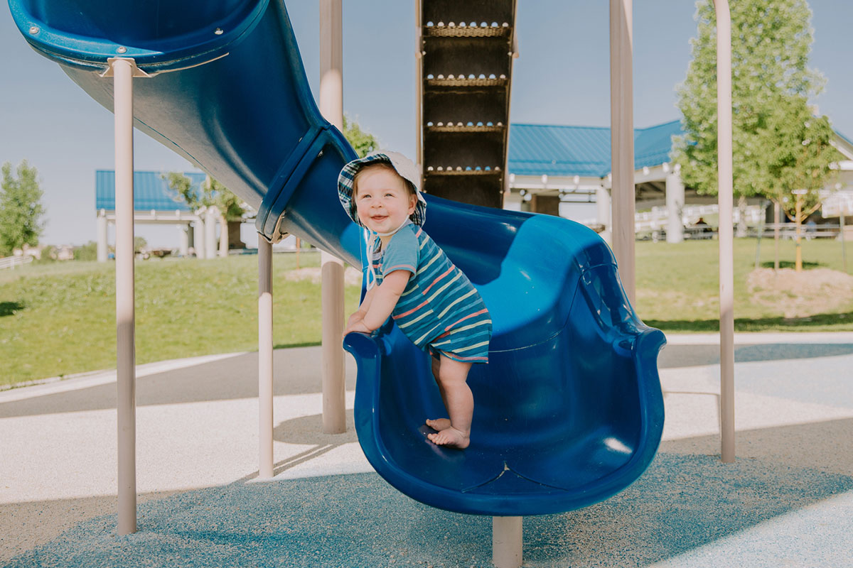 Smiling toddler in a striped outfit and sun hat standing at the end of a blue playground slide in a sunny park.