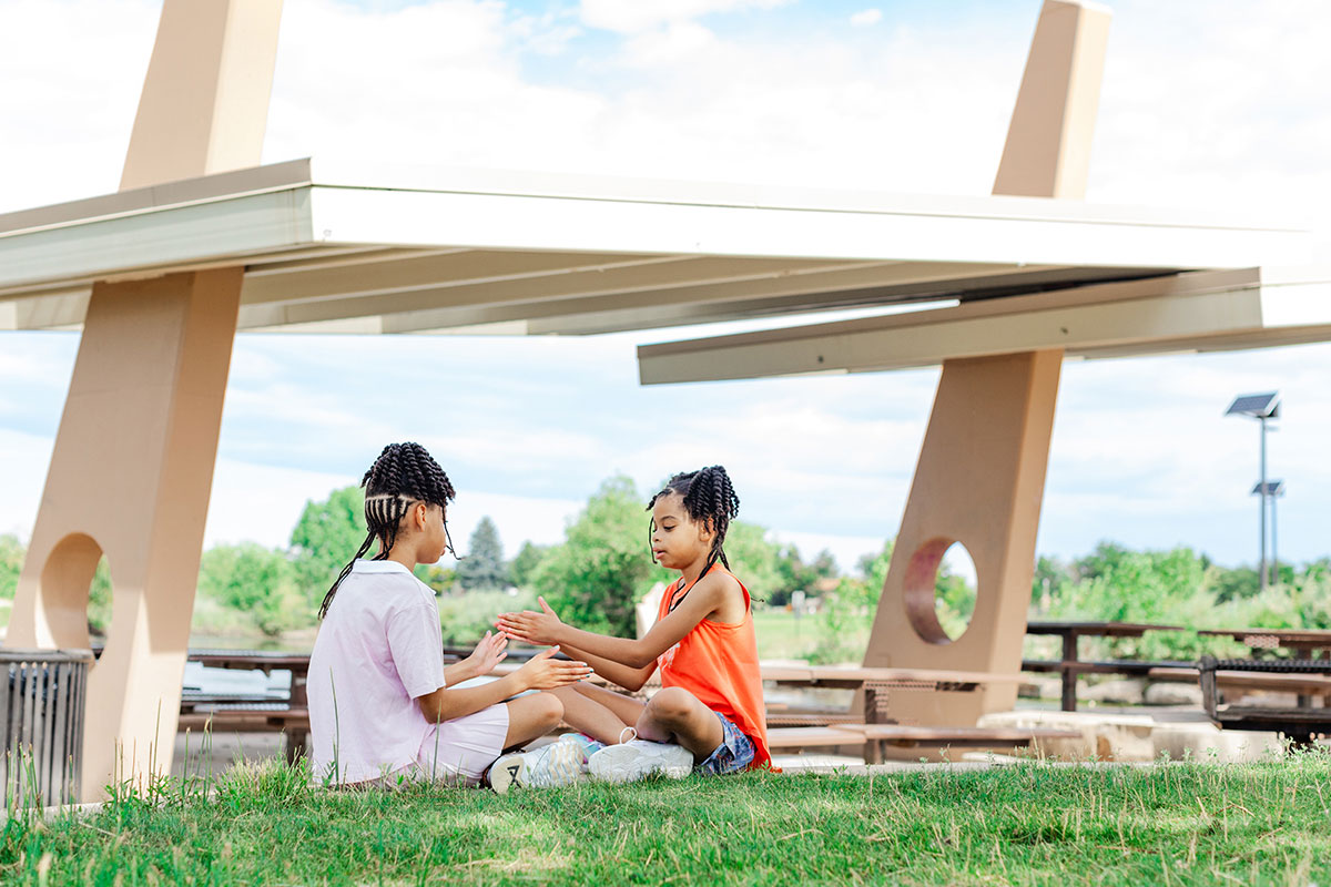 Two children sit on the grass playing a hand-clapping game in a park. 
