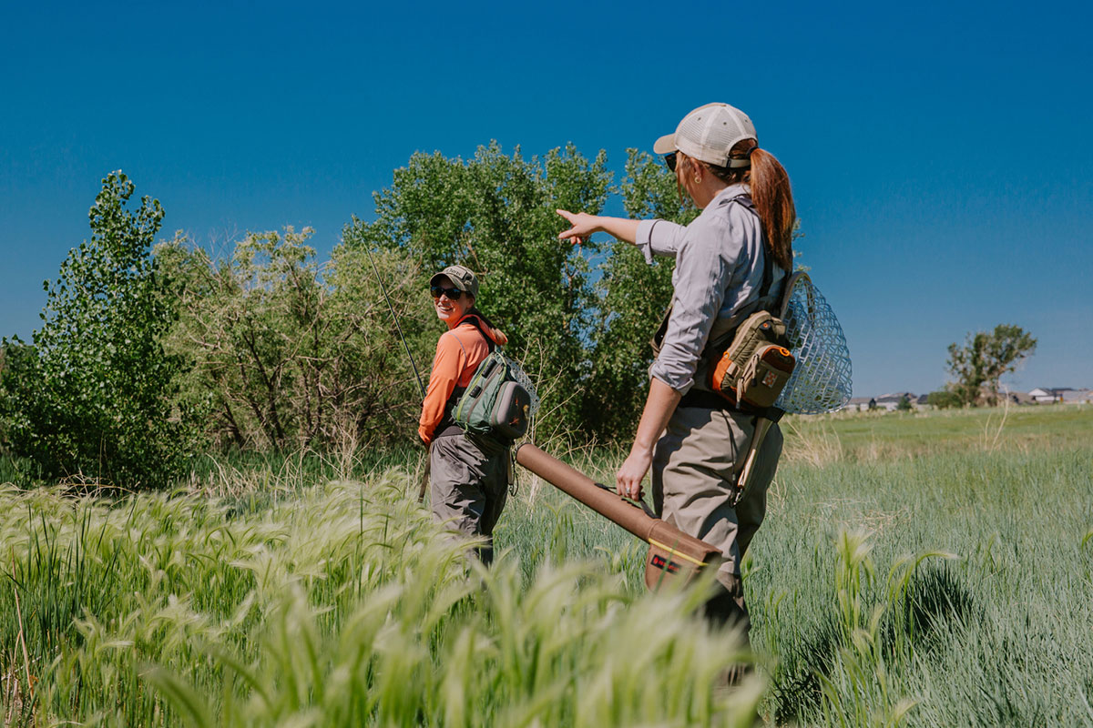 Two people in waders carry fishing gear and walk through tall grass in Aurora, Colorado