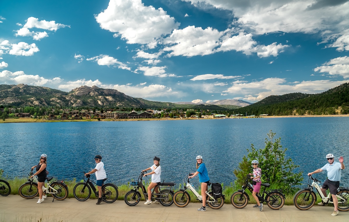 E-bike riders smiling at the camera on a path with water and hills in the background in Estes Park, Colorado.