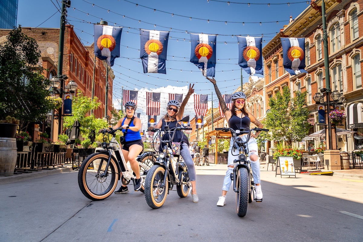Three women on e-bikes posing under Colorado flags in Denver, Colorado during a Ride Colorado tour.