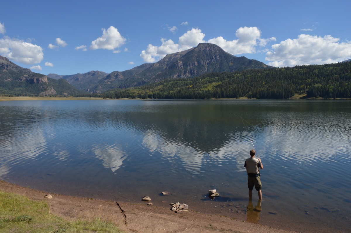 A man fishes beside calm, glassy waters with scenic alpine scenery in Pagosa Springs, Colorado