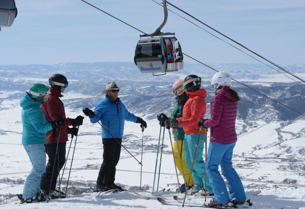 Five skiers in colorful gear listen to another skier at Steamboat. In the background a gondola makes it's way up the mountain on a winter's day. The ground beneath them is covered in snow.