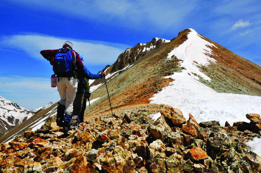 A mountain climber stands on rocky ground and stares up at a mountain peak that's covered in snow. There's a blue sky with slight white clouds. 