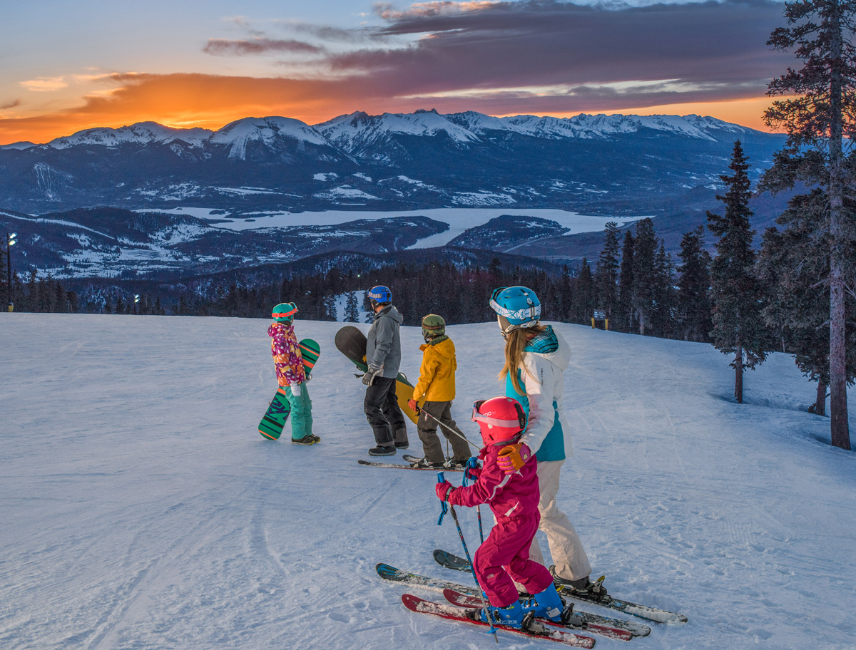 Two adults and three kids, all equipped for snowboarding and skiing, look off into the distance as the sky behind the distant snow-capped mountains turns a vibrant golden-orange at a ski resort in Colorado.