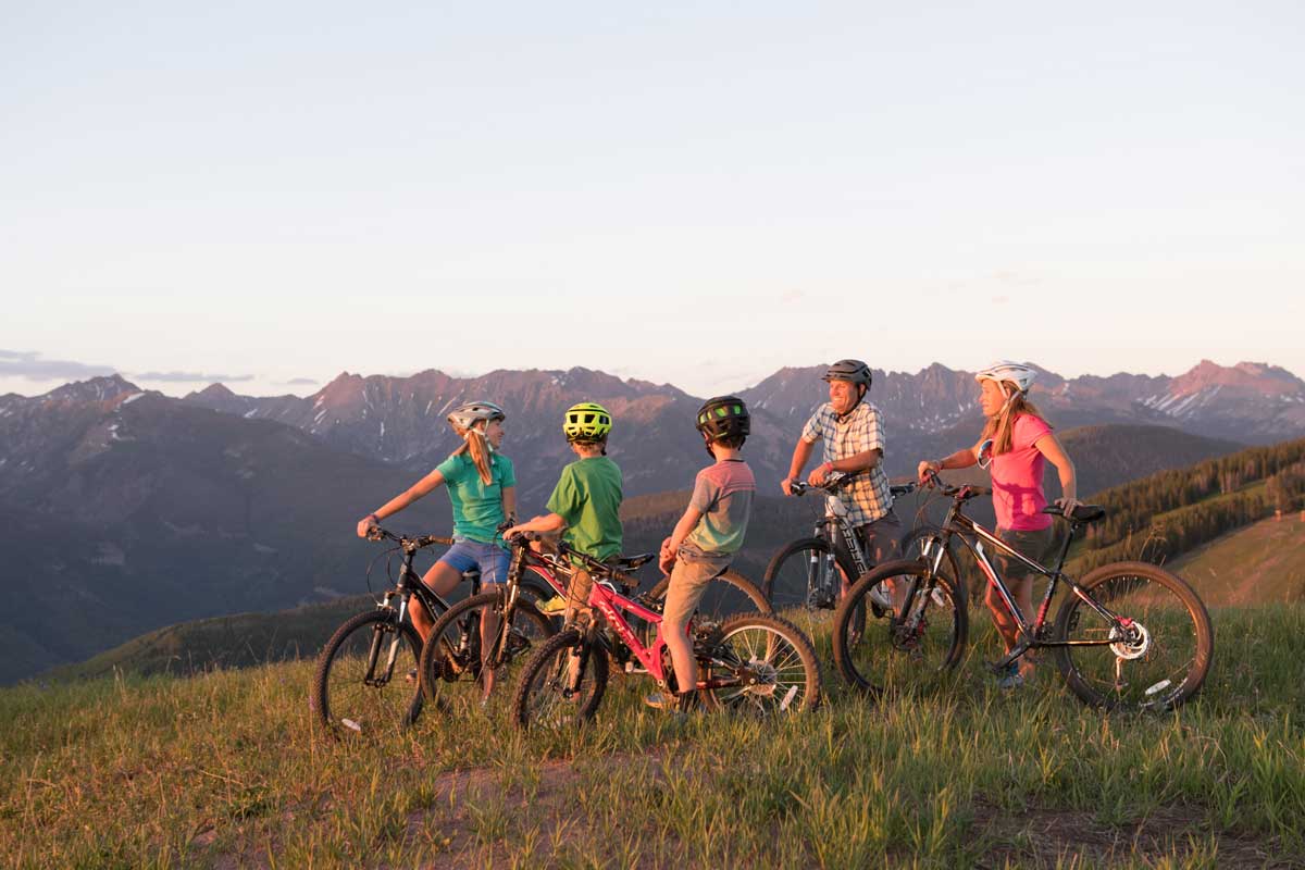 Two adults and three kids pause with their mountain bikes atop a grassy ridge on Vail Mountain. The sun is setting and paints the people and mountains in a pinkish glow.