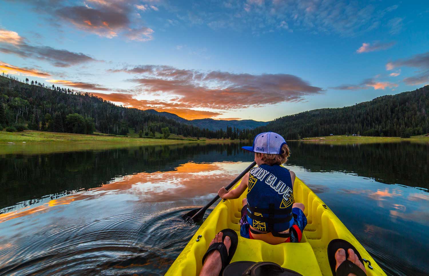 A boy in a yellow kayak and a life vest paddles through still waters toward an evergreen-lined shore as the sun sets