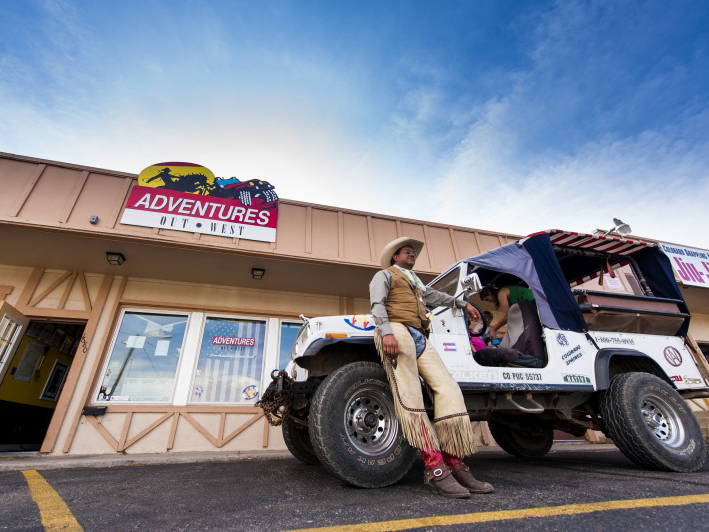 A cowpoke dressed in fringed chaps and a leather vest leans against small white jeep in Colorado Springs, Colorado.