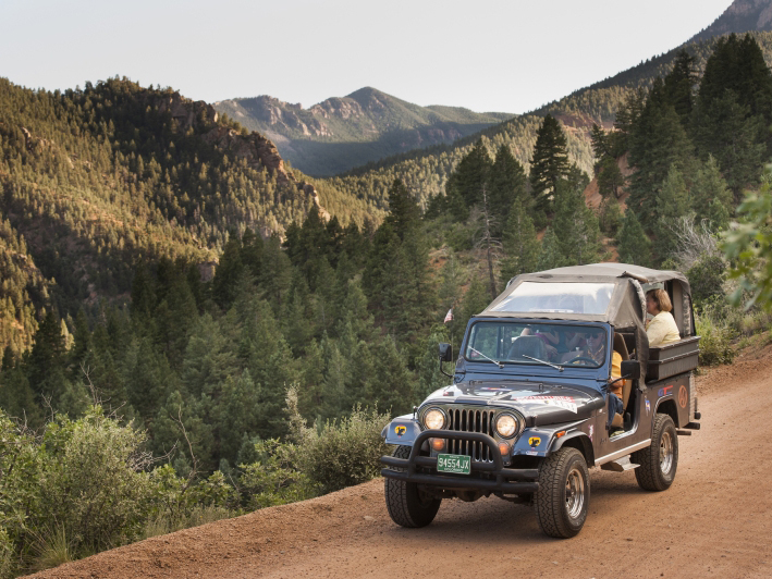 A tour group rides in a covered blue jeep down a dirt road in Colorado. A pine forest stretches out in the valley beside the road.