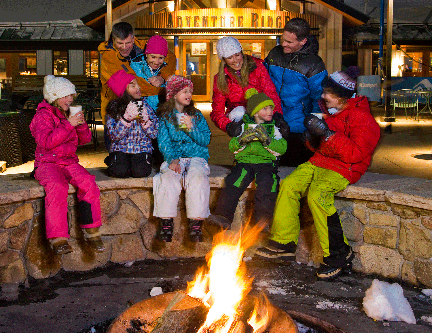 A group of kids and adults dressed in ski clothes sip hot chocolate around a crackling firepit in a ski village