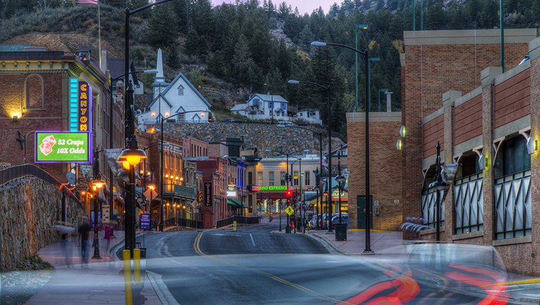 Downtown Black Hawk at sunrise. The street is empty of cars and the neon signs are ablaze. The brick buildings that sit along the winding street are all two to three stories. Up on a hill above the street sits a white church and house. There are evergreen trees above that.