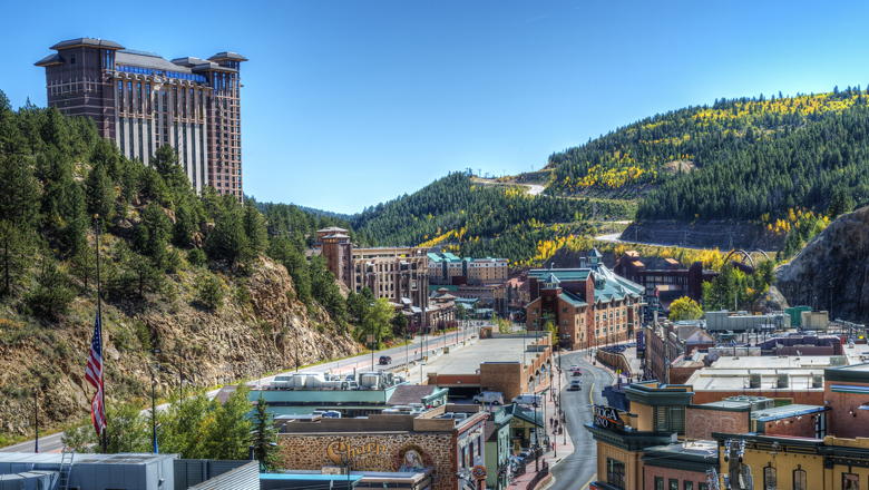 Downtown Black Hawk on a blue-sky fall day. In the distance aspen trees are starting to turn golden, and a winding road is making its way up a mountain. The winding Main Street has a couple cars driving by historic brick buildings. On the left a hulking building is being built above town. 