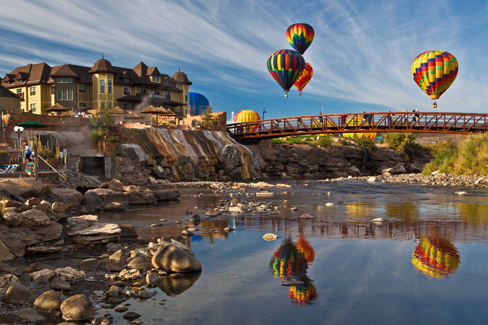 Four hot air balloons fly above a glassy river with a metal pedestrian bridge in the distance. On the right a building with a rocky riverbank.