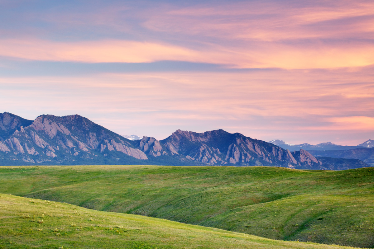 Green rolling hills stretch towards Boulder's slanted Flatirons rock formations. The clouds above are tinged pink and purple.