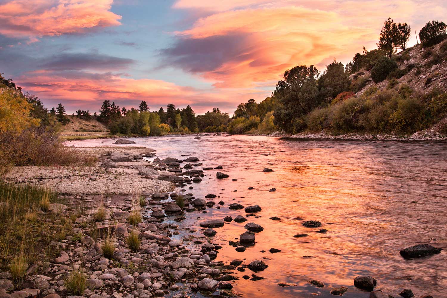 A colorful sunset reflects on a still river surrounded by mountains 