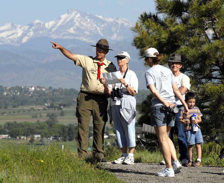A park ranger points to the right showing a family of four, three adults and one child where to go in the summer with green grass and trees. In the background a hazy snow-capped mountain sits under a blue sky.