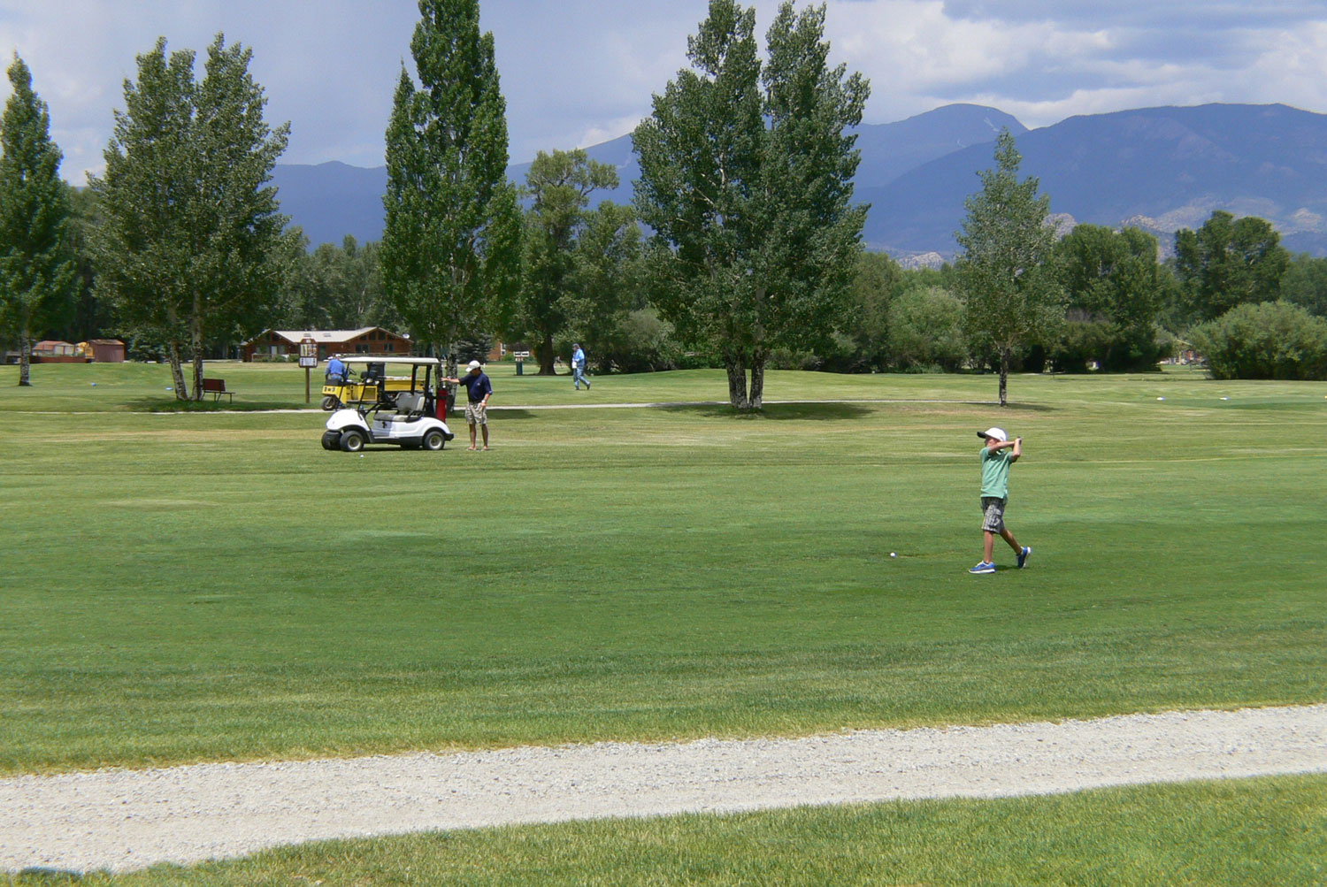 A young man takes a big swing while his golf partners watch from a golf cart. The grass is green, the trees are green and there are rolling blue hills in the background