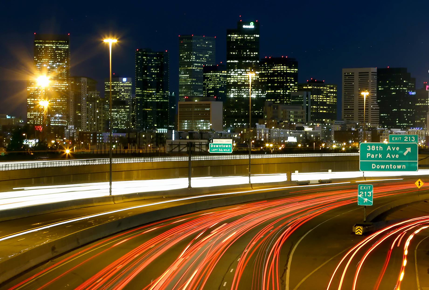 The skyscrapers of a Colorado city are lit and stand tall against a dark, night sky. In front of the city are curving highway road with green exits signs.