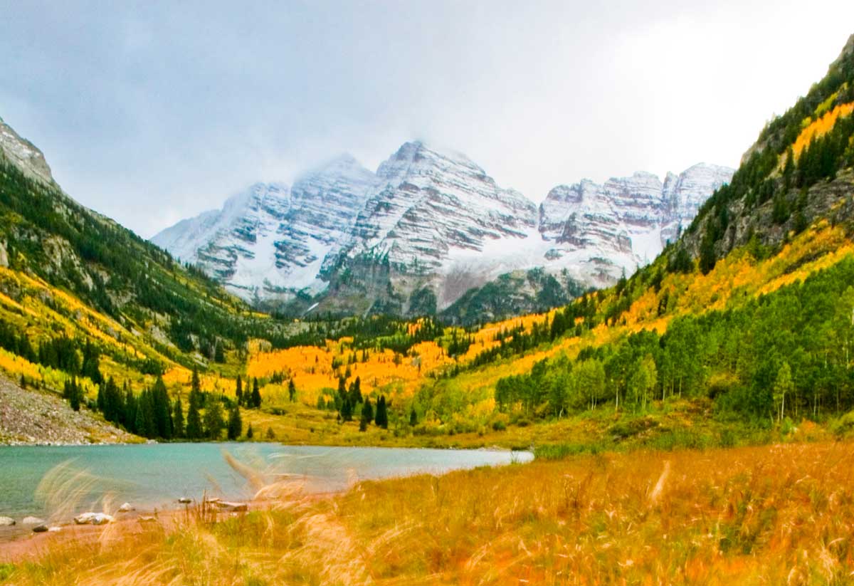 The snow-covered Maroon Bells near Aspen & Snowmass sit above a valley of golden aspens and evergreen trees near a mountain lake.