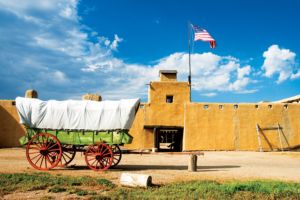 A green-painted wooden wagon with red, spoked wheels and a white canvas top stands outside an old fort near Bent, Colorado.