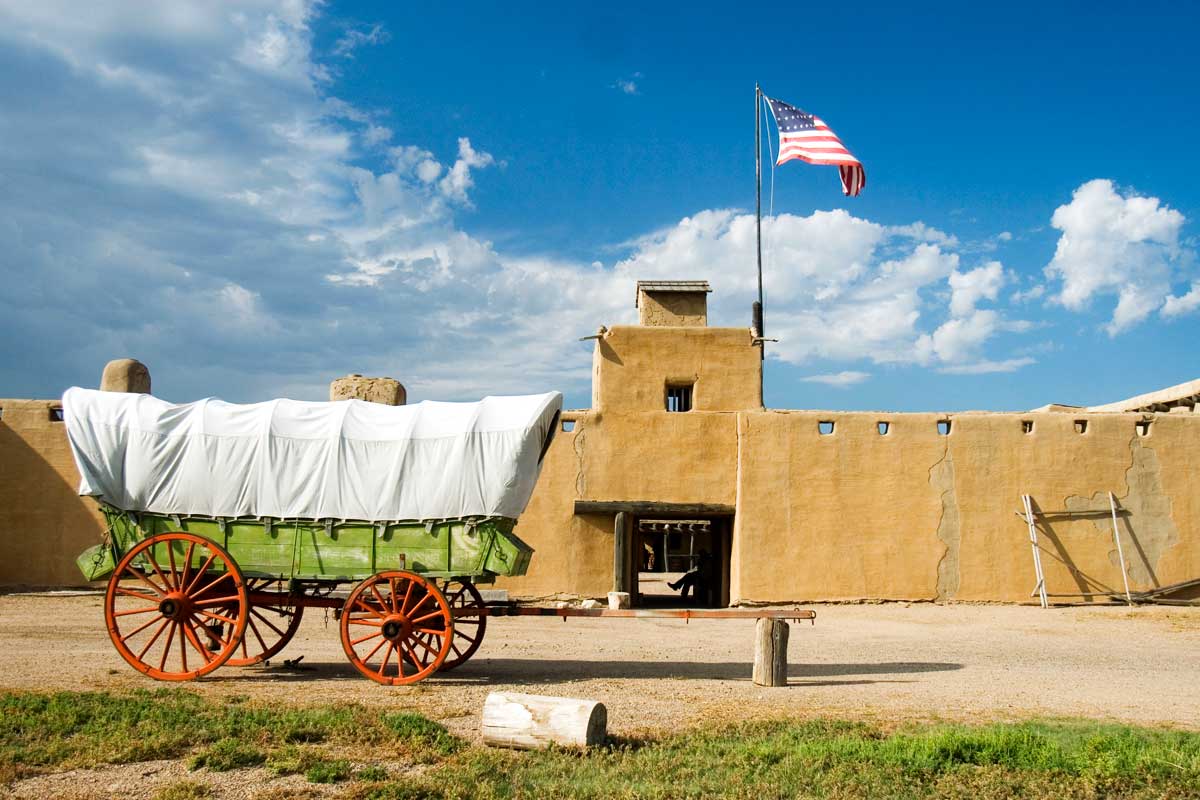 A green wagon with a white cover sits in front of the recreated Spanish-style Bent's Old Fort National Historic Site on a blue-sky day with white clouds.