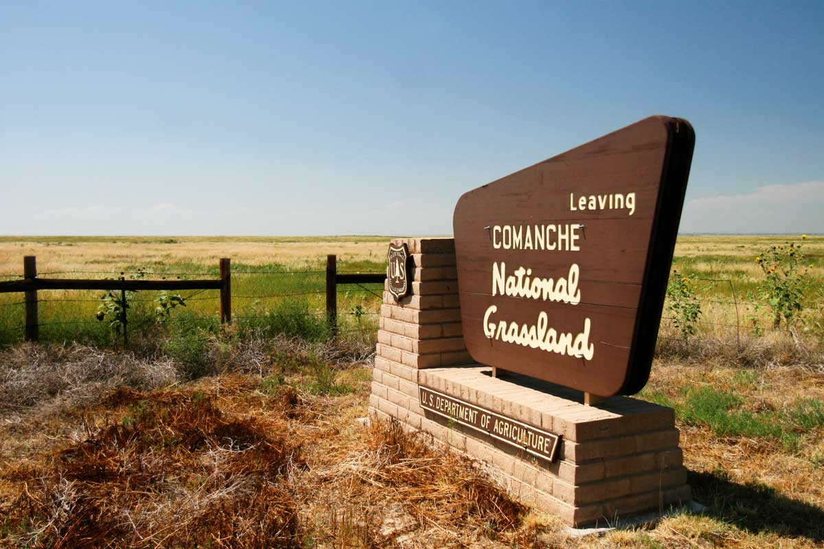 A blue sky with a haze that has a green field with a wooden fence sits behind a "Comanche National Grassland" brown sign.