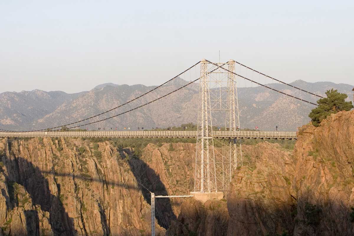 The metal Royal Gorge Bridge rises above the stone canyon face with Rocky Mountains in the distance under a hazy sky.