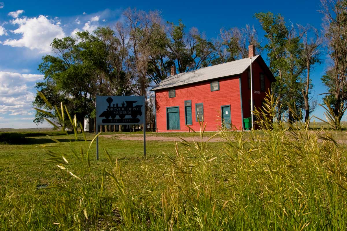 A red wooden building sits on green grass with green-leafed trees in the background at Grover Depot Museum