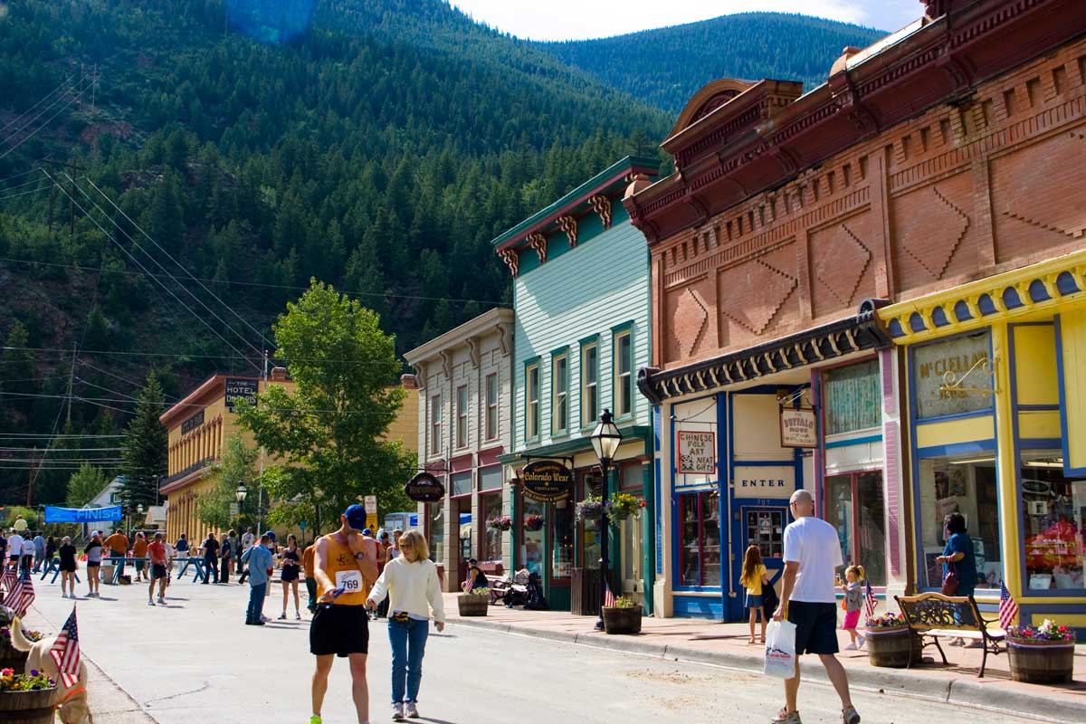 People walk down a street in Georgetown. On the right of the image are historic Victorian storefronts. In the distance evergreen-covered mountains sit.