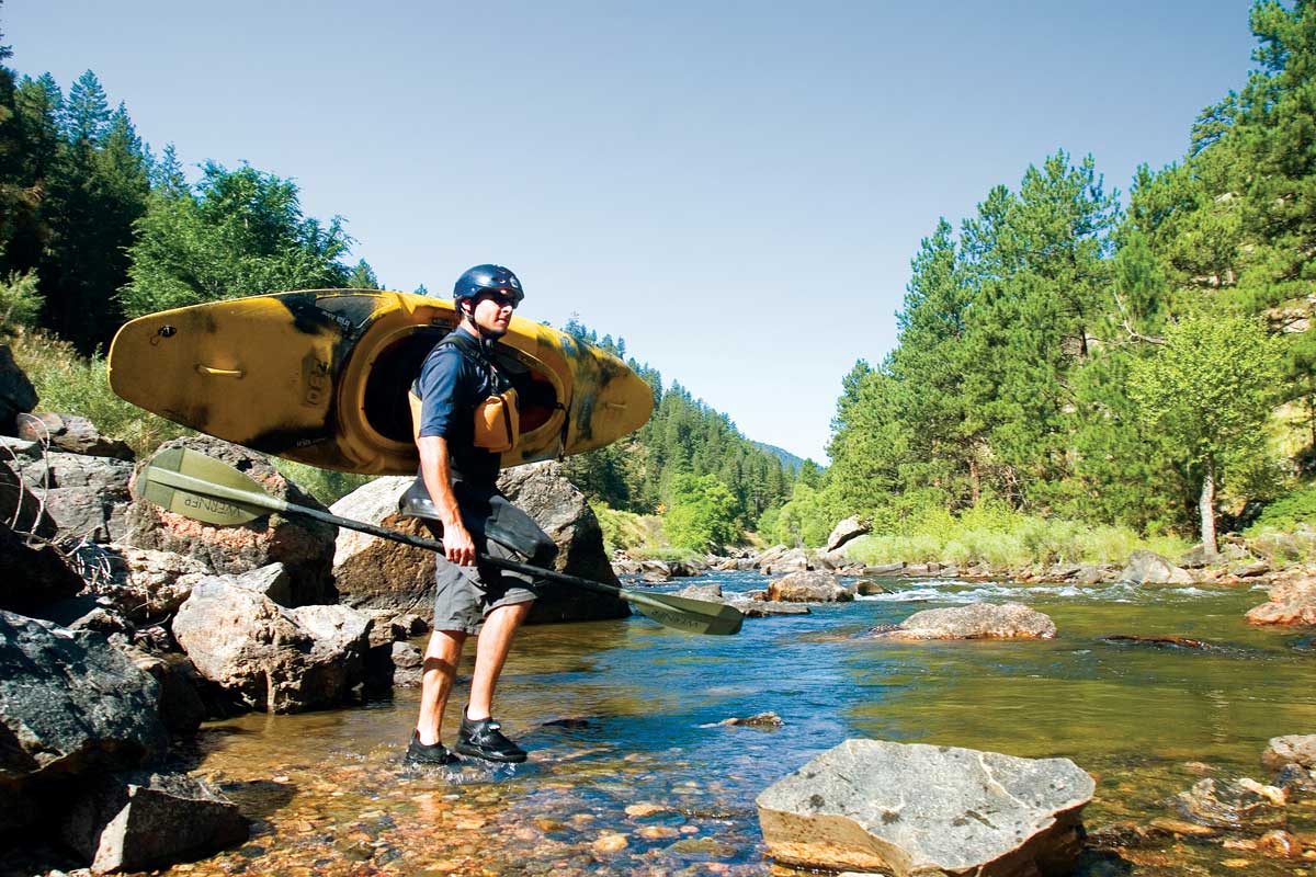 A kayaker wearing a bright yellow life vest carries a yellow and dark blue kayak with one arm by the shallows of the Cache la Poudre Wilderness near Fort Collins, Colorado.