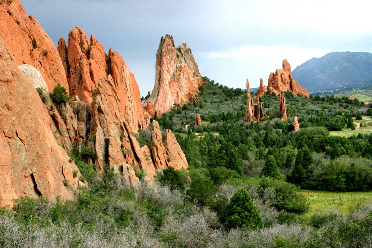 On the left side, thin red-rock formations protrude from the earth. On the right green-grass fields lead up to dark-green bushes at the base of the geologic formations at Garden of the Gods.
