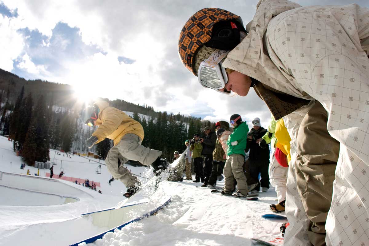 Snowboarders stand on the edge of a bowl at the Terrain Park at Vail Resort. A snowboarder in a yellow jacket is in the air as they begin their dissent.