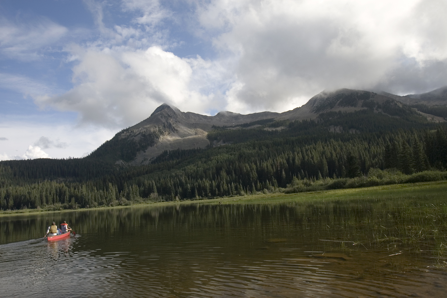 A red canoe with two people in it sits in the middle of a calm lake with surrounded by evergreen trees. There's a stone-mountain peak in the distance.