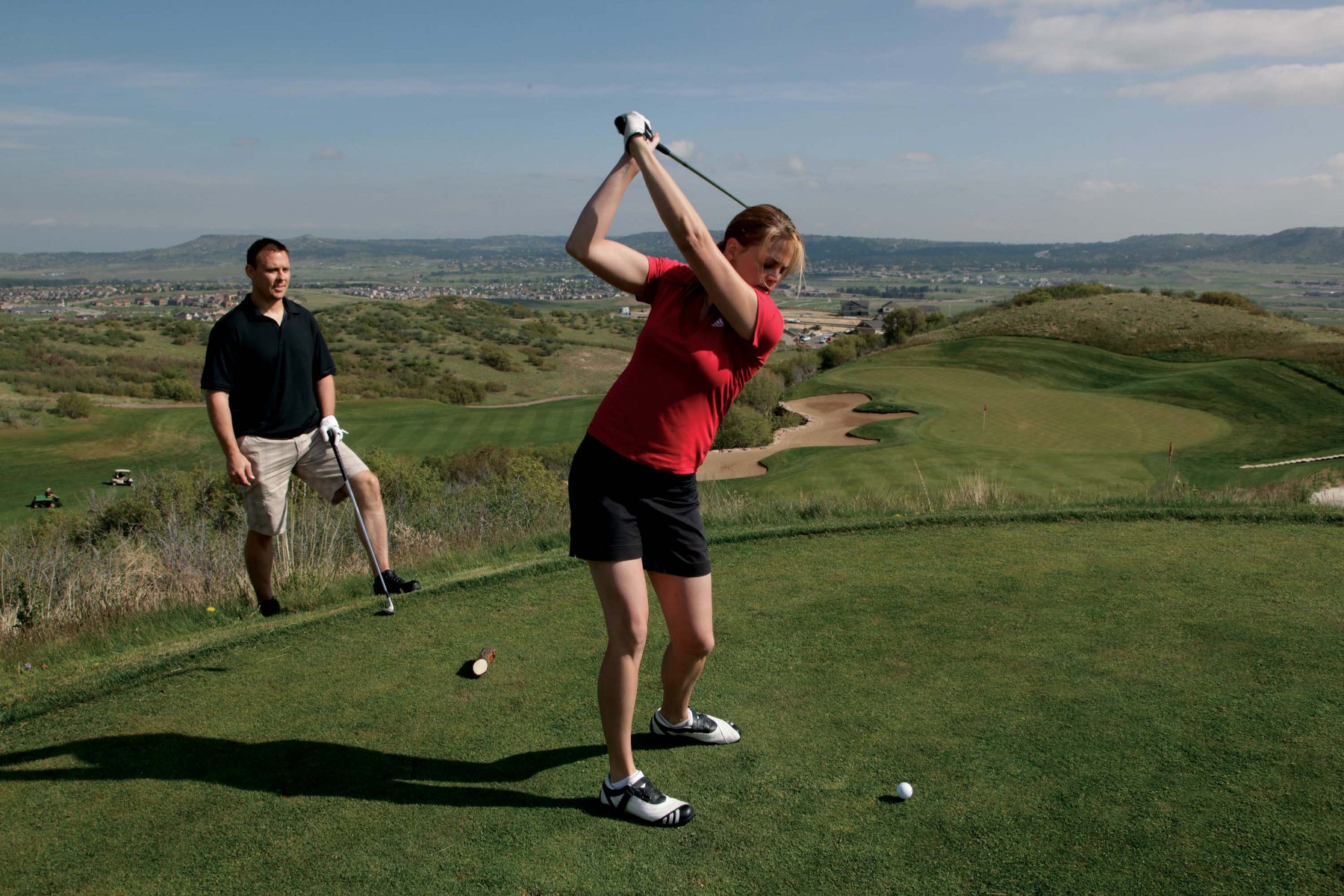 A woman swings her golf club as a man watches on, on a green golf course with a hazy blue sky and a town in the background.