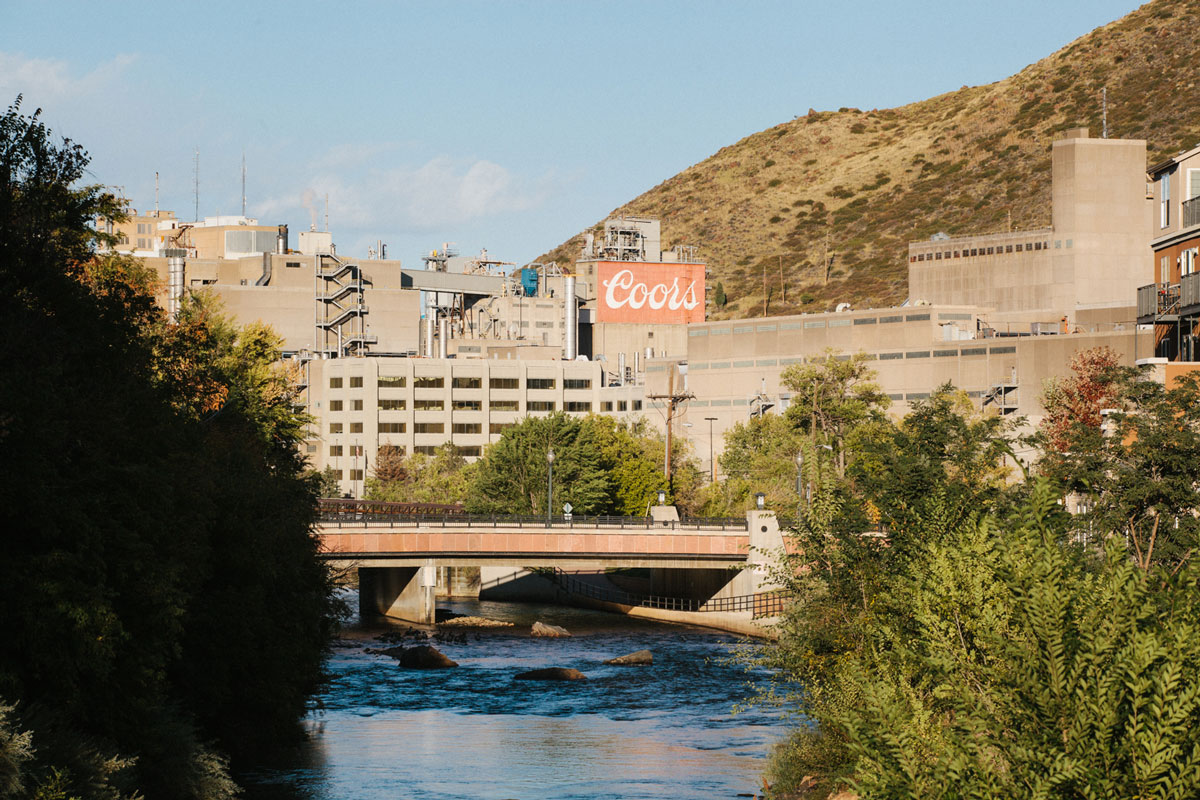 The gray-building conglomerate of Coors Brewing sits surrounded by green trees behind a blue river with rolling hills on the right.