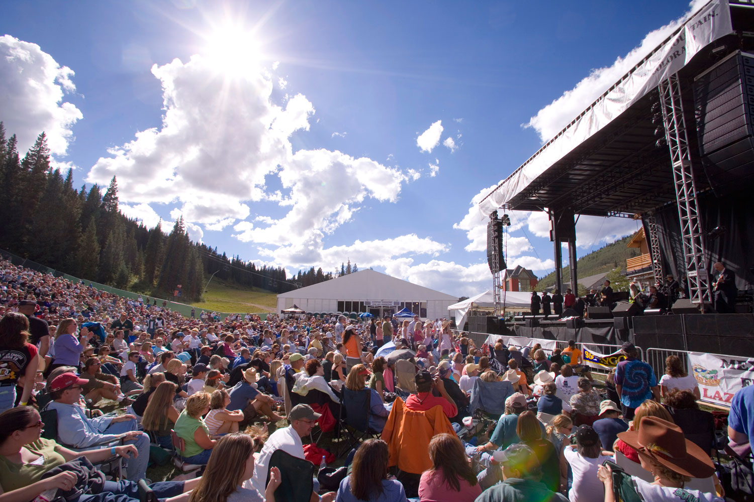 An outdoor concert stage with a big crowd in front of it on a summer's, blue-sky day with evergreen trees at Copper Mountain.