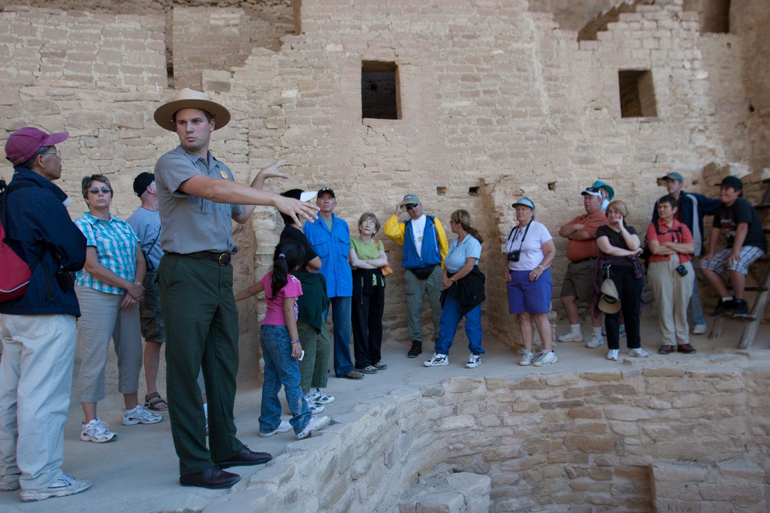 A ranger dressed in dark green pants, a khaki shirt and a wide-brimmed hat stands in front of an open pit and speaks to a tour group at Mesa Verde National Park.