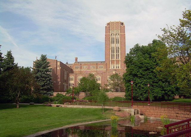 Across a grassy quad, a sandstone building with a tower peaks out from behind several trees at the University of Denver campus.