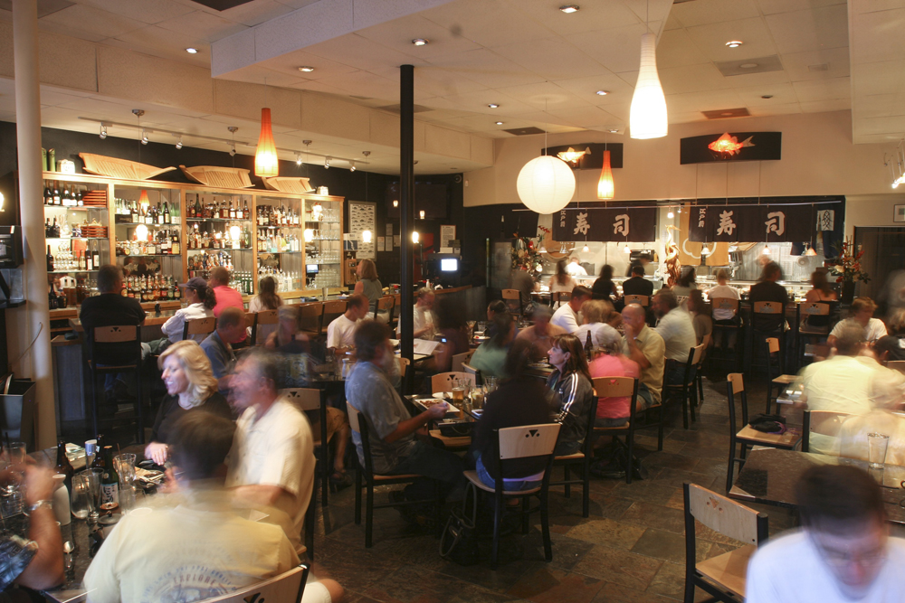 The crowded dining room of a restaurant; the bar is stocked with bottles and you can see chefs working in the kitchen beneath banners with Japanese characters on them