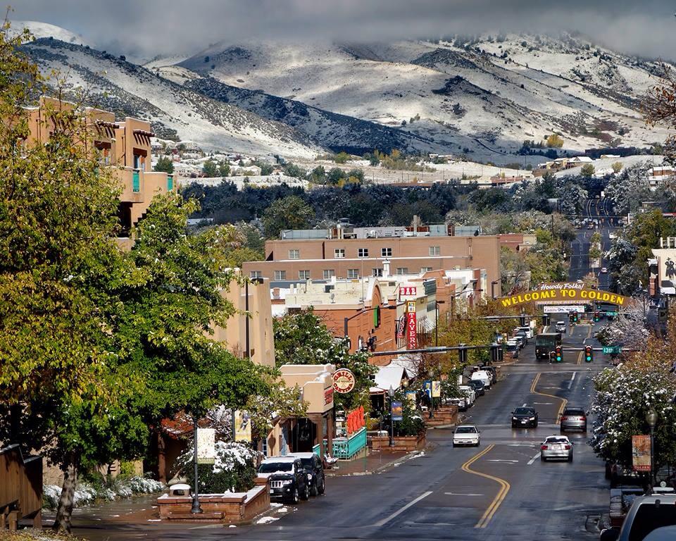 A view of downtown Golden with snow-covered foothills in the distance. The arched "Welcome to Golden" sign sits at the bottom of the photo with cars driving both directions on the road and green trees intermixed with historic and new buildings.