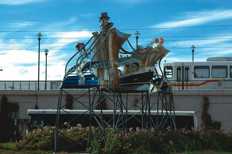 Against a blue sky, with a light rail train in the background, the Red Grooms sculpture sits tall. Made out of metal it depicts a man who is also a bridge.