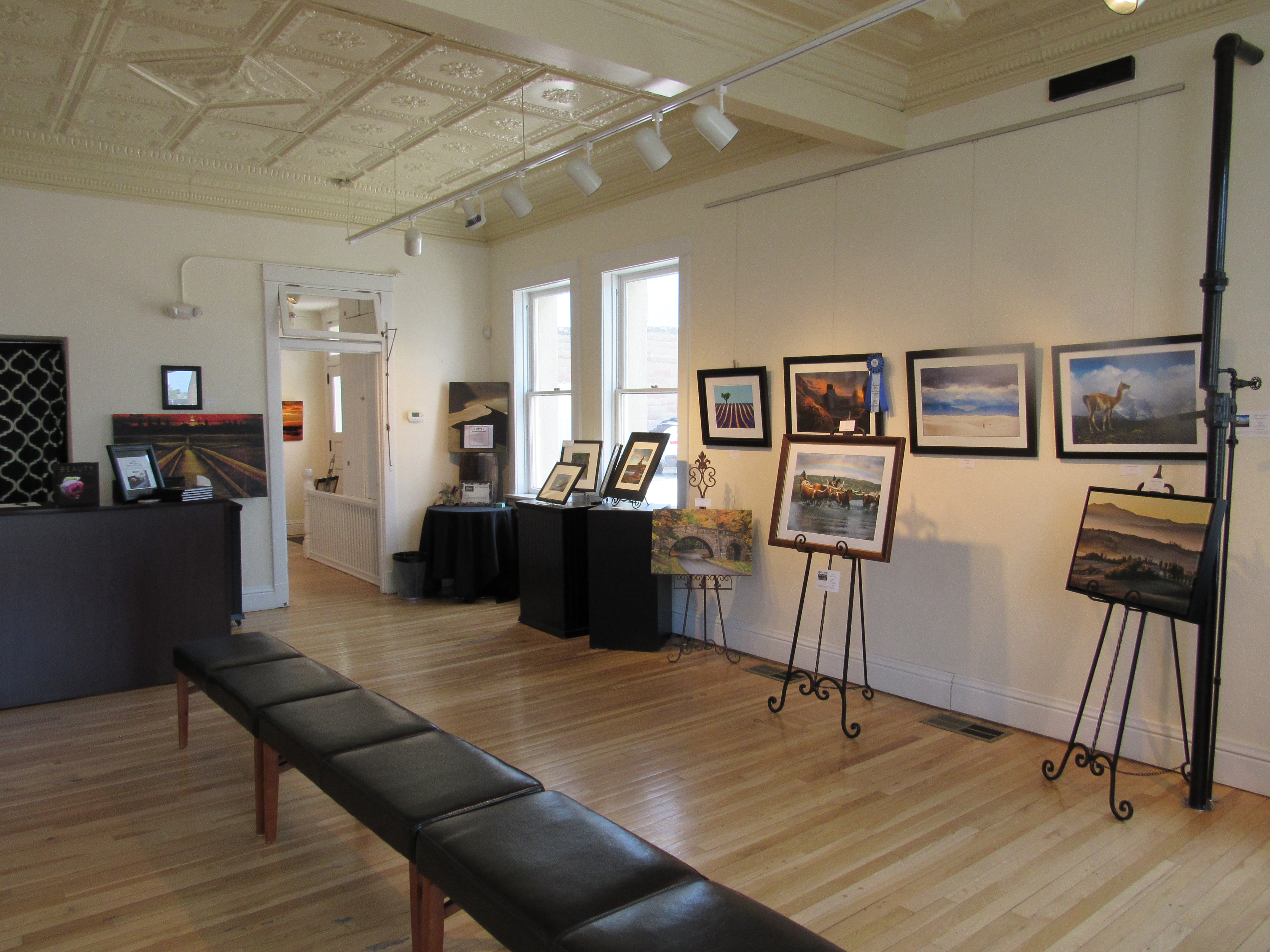 An exhibition at Windsor Art & Heritage Center is on display in a room with white walls and light-wood floors. There's a leather bench sitting in front of easels with art on them.