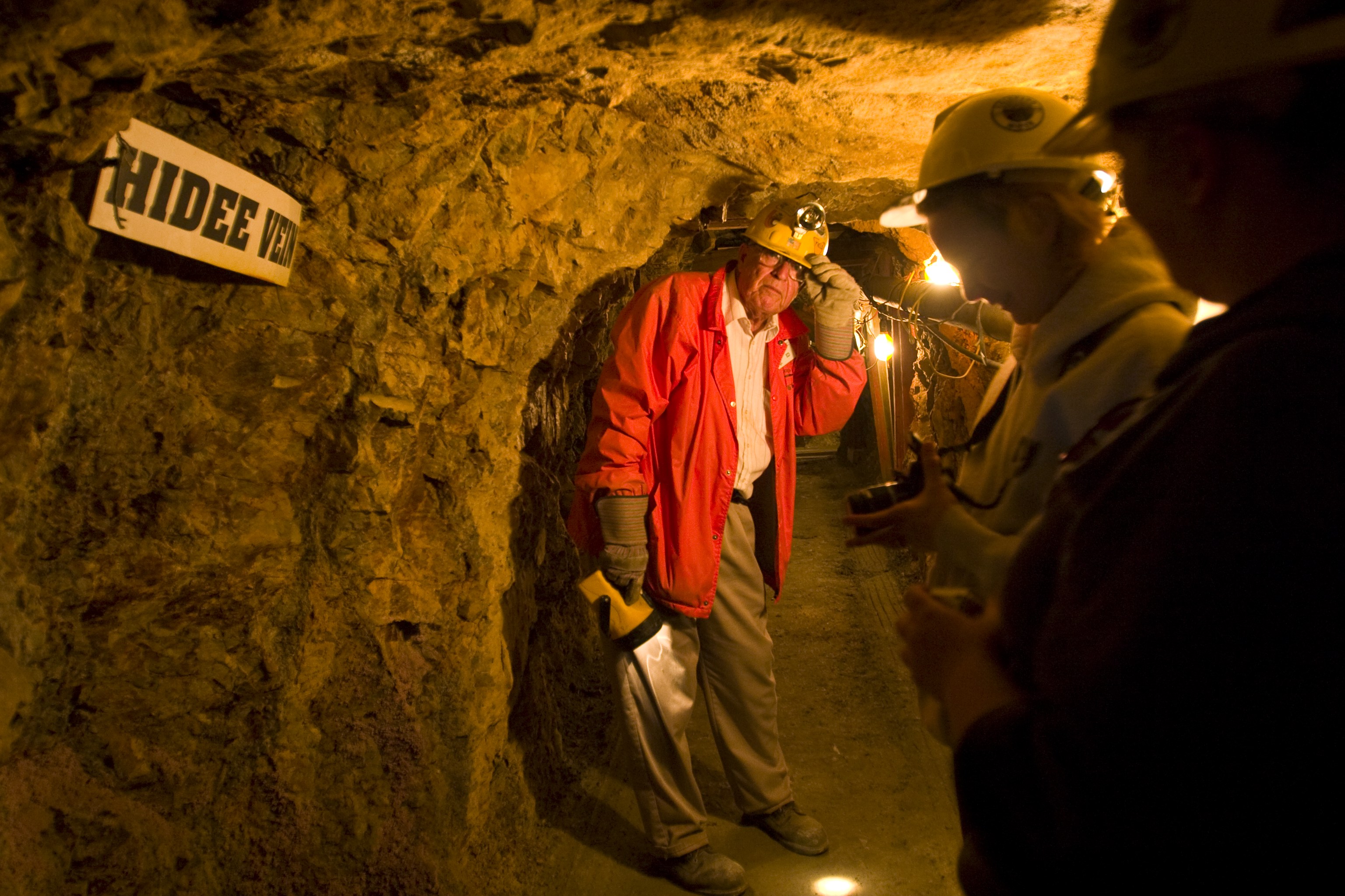 A tour guide in a red jacket and yellow hard hat leads a group of people through a dimly lit tunnel