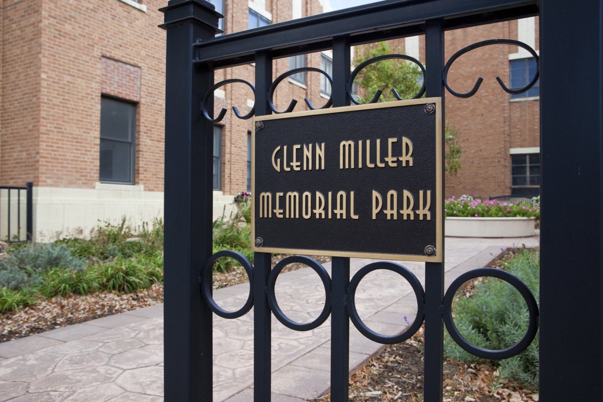 A wrought-iron gate with a sign rimmed in gold, with gold letters that say "Glenn Miller Memorial Park" sits slightly ajar to a sidewalk with green plants on each side. There are red-brick buildings in the distance.