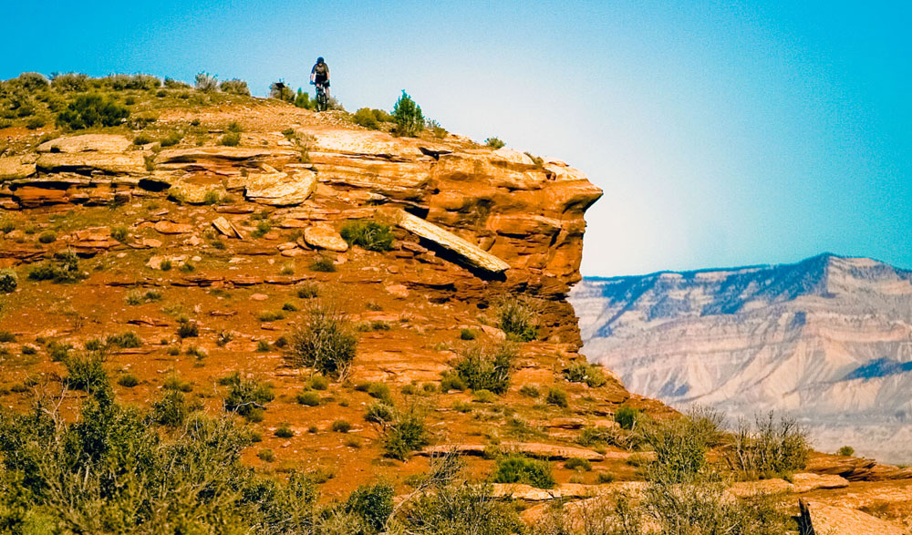 Way atop a giant orange-colored hill with splotches of sagebrush, we see a wee mountain biker making their way along a trail