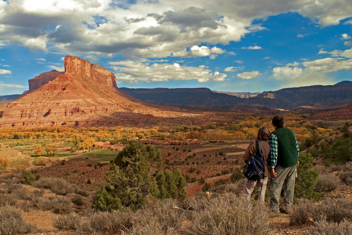 Two hikers stand with their backs to the camera on the right side staring at a red-rock butte in the distance. Between them and the formation is a valley with trees and red-dirt. There's a blue sky and white clouds.