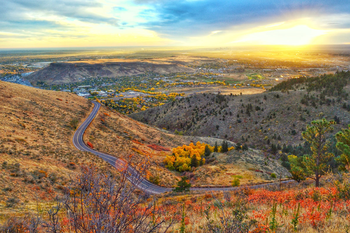 Atop a mountain, a winding road leads through yellow grasses with golden trees intermixed with evergreens overlooking downtown golden at sunrise. The sky is yellow with blue clouds. 