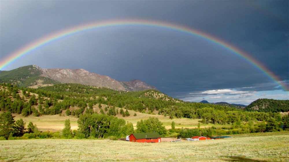 A dark stormy sky with a giant rainbow sits above a Rocky Mountain range with green trees and below that a valley of grasses and red barns.